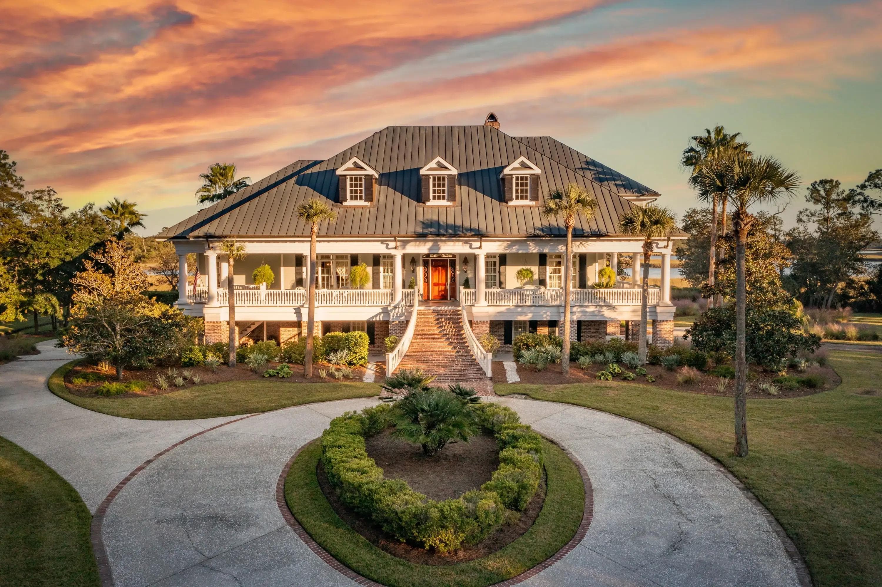Stately white mansion with a metal roof, grand staircase, and circular driveway at sunset.